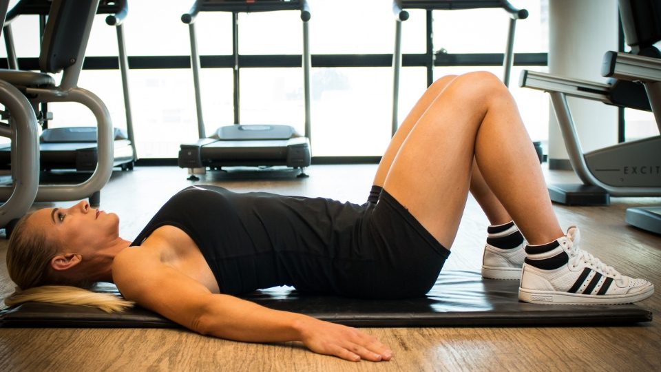 A woman in a gym lays on a mat with bent knees, wearing black athletic wear and white sneakers. Treadmills are visible in the background, conveying focus and determination.